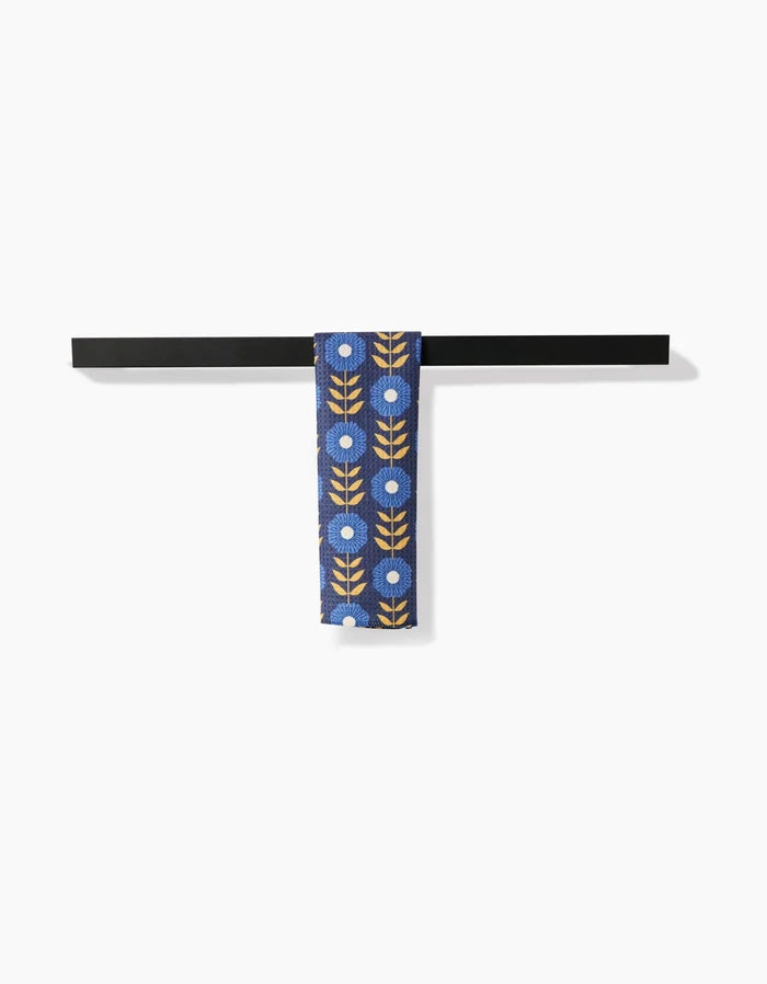 The Scandi Floral necktie by Megan Kampa, crafted from eco-friendly Geoweave fabric, displays white flowers and yellow leaves on blue, hanging over a black bar with a white background.