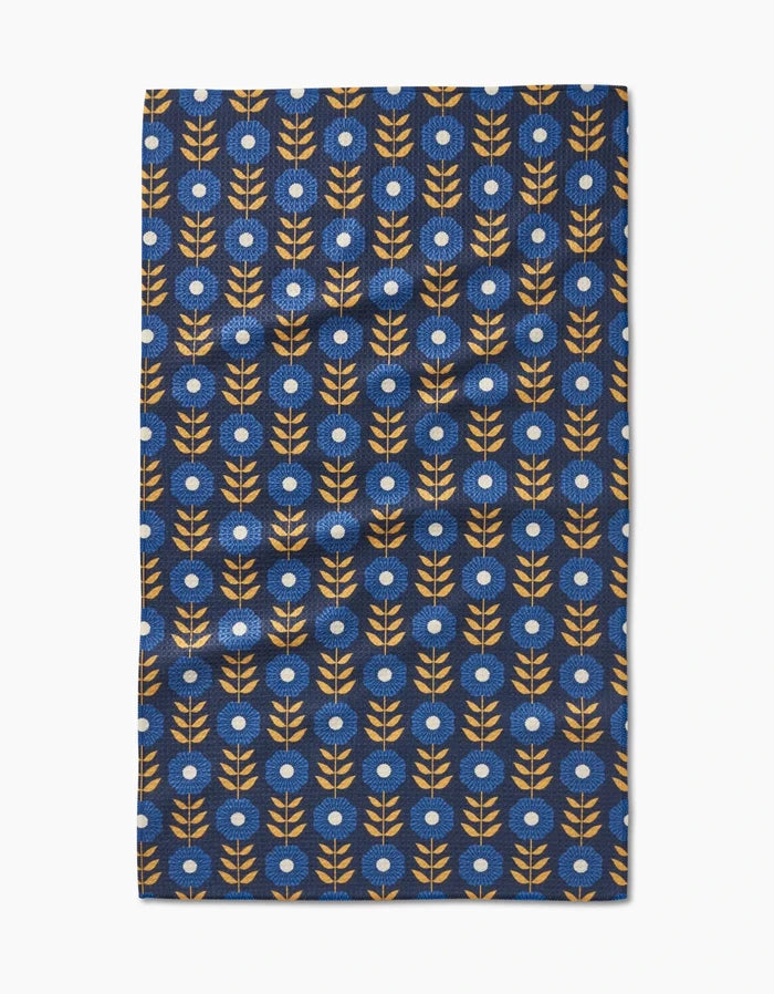 The Scandi Floral tea towel by Megan Kampa features stylized orange branches and white-centered blue flowers on dark blue Geoweave fabric, blending geometric beauty with practical function.