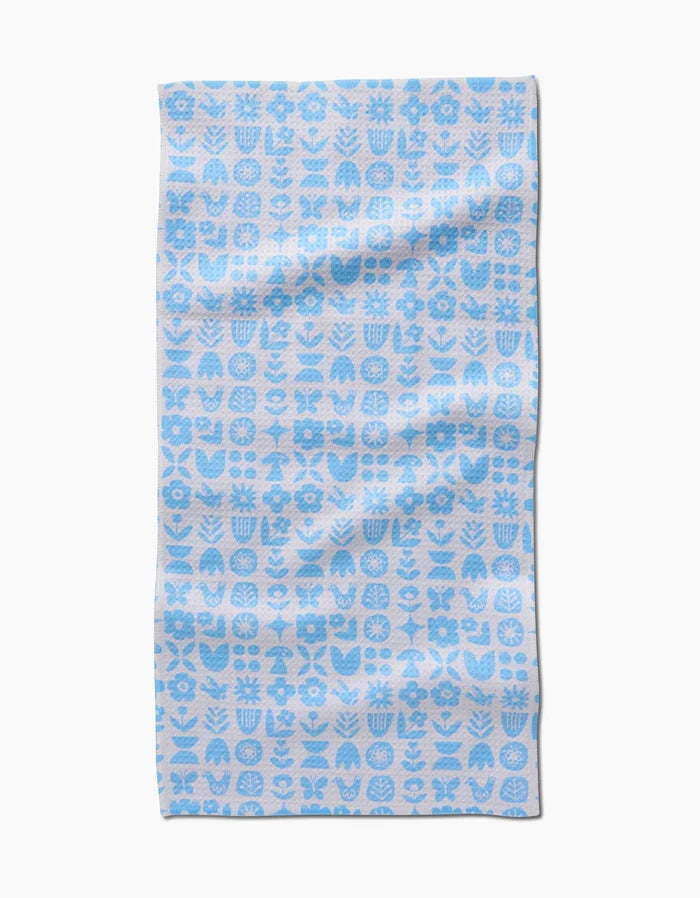 The Scandi Spring Blue Bar towel by Kayla Ann features a blue and white grid pattern of stylized plants, flowers, and geometric shapes. Made from Geoweave fabric, it is shown slightly wrinkled and lying flat on a white background.