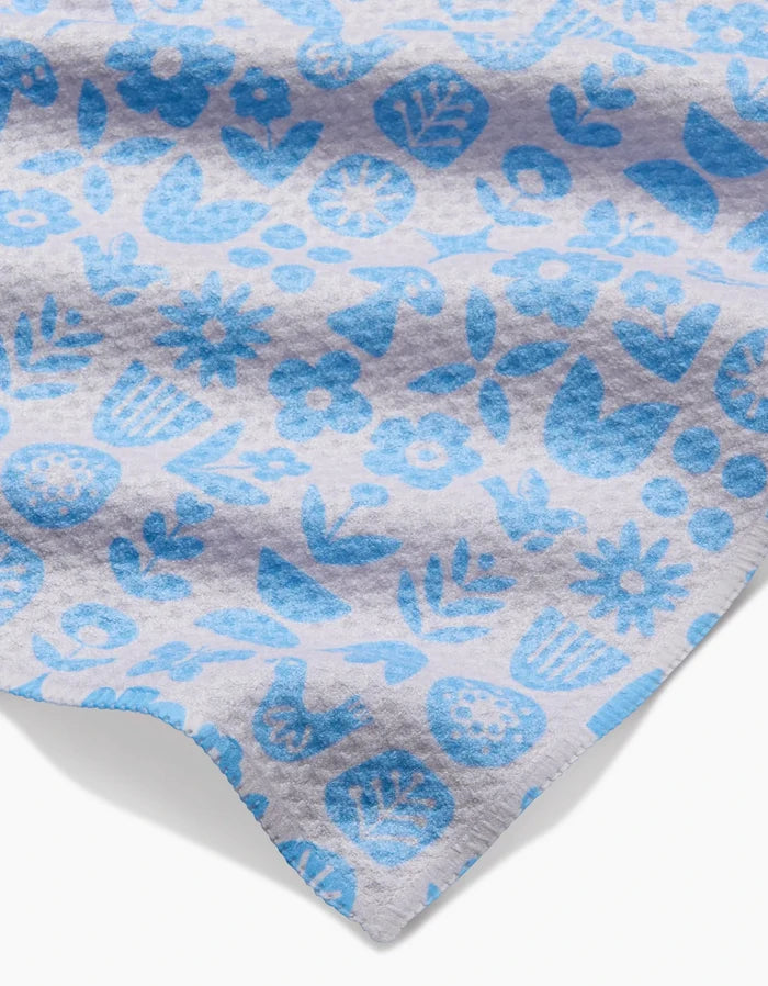 A close-up of the Kayla Ann Scandi Spring Blue bar towel shows its light gray Geoweave fabric with a blue floral and leaf pattern. Made from post-consumer recycled materials, it has a soft texture and slightly wavy edge.