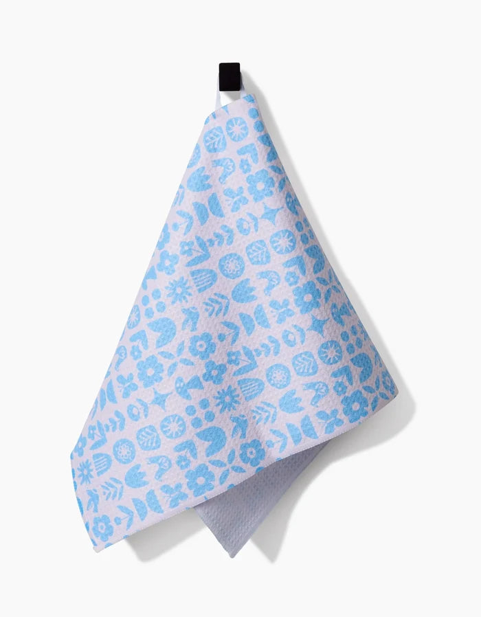 The Scandi Spring Blue bar towel by Kayla Ann features a floral and leaf pattern in light blue, made from post-consumer recycled materials, and hangs from a black loop against a white background.