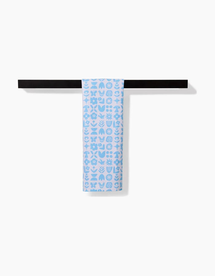 The Scandi Spring Blue bar towel by Kayla Ann, featuring white geometric and botanical patterns on light blue Geoweave fabric, hangs neatly over a black towel bar against a plain white background.