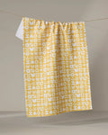 The Kayla Ann "Scandi Spring" yellow tea towel, made from recycled materials and adorned with a playful white floral pattern, hangs over a clothesline against a neutral background.