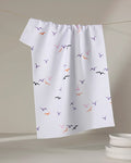 The "Scattered Seagulls" tea towel by Ania Zwara Art, featuring vibrant bird silhouettes, hangs on a line beside a stack of white dishes on a light surface.