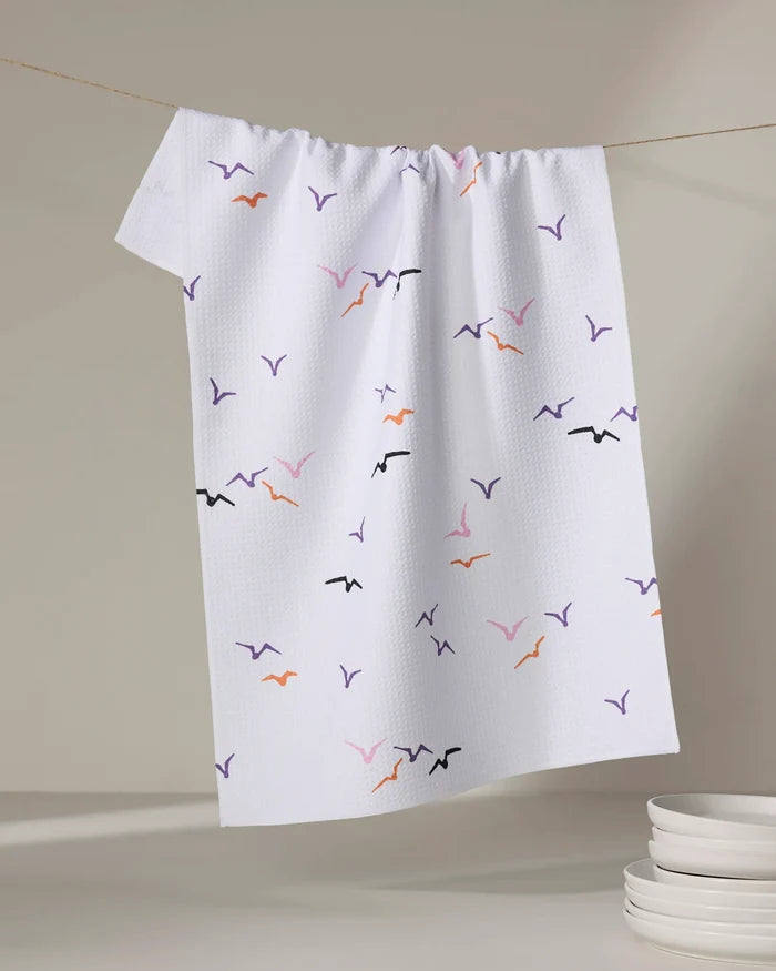 The "Scattered Seagulls" tea towel by Ania Zwara Art, featuring vibrant bird silhouettes, hangs on a line beside a stack of white dishes on a light surface.