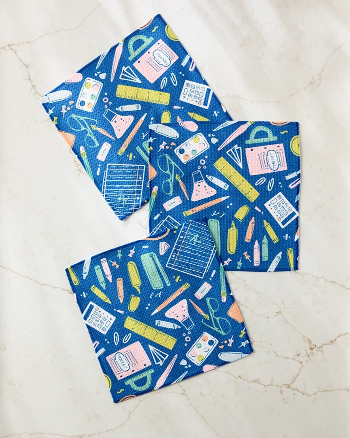 The "School Days" dishcloth set by Amy MacCready features three blue cloths with vibrant school supplies patterns. Made from recycled materials, they're perfect for eco-friendly cleaning and look stylish on any white marble surface.