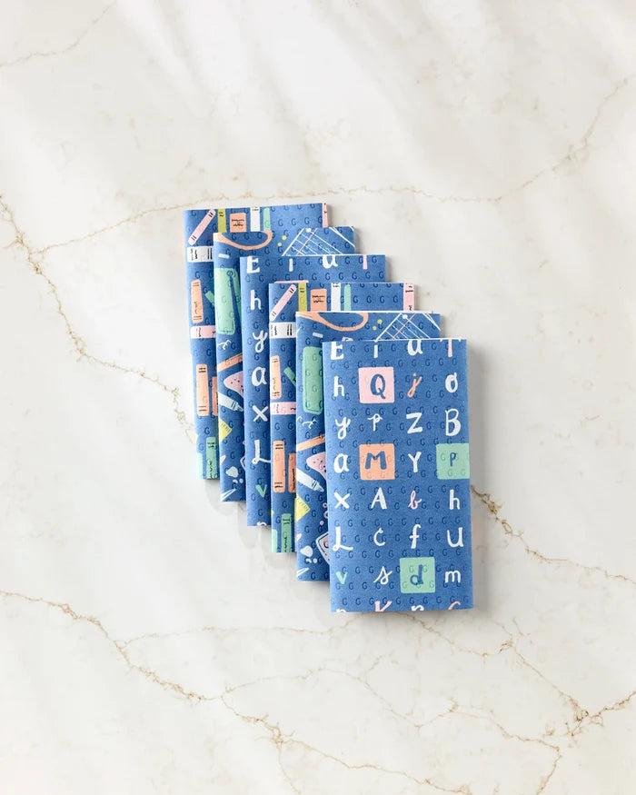 Seven "School Days" blue notebooks by Amy MacCready, featuring alphabet and geometric patterns, are stacked on a marble surface—crafted from post-consumer recycled materials for eco-friendly use.