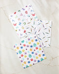 Three Scribble Squad absorbent kitchen towels by Vaishali Design Studio are displayed on a light marble surface: one with playful letters and animals, another with scattered crayons, and the third featuring cheerful cartoon monsters.