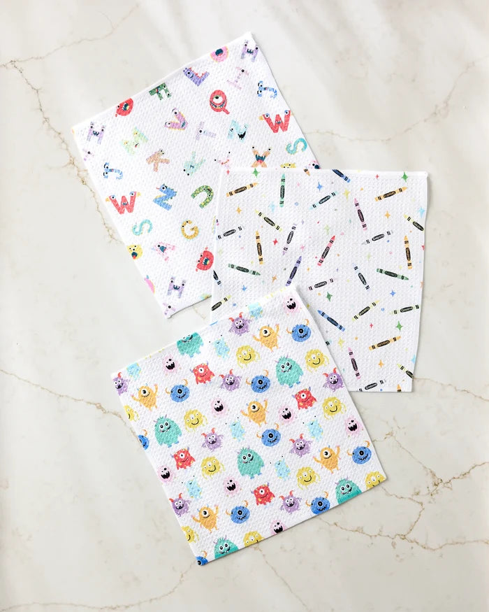 Three Scribble Squad absorbent kitchen towels by Vaishali Design Studio are displayed on a light marble surface: one with playful letters and animals, another with scattered crayons, and the third featuring cheerful cartoon monsters.
