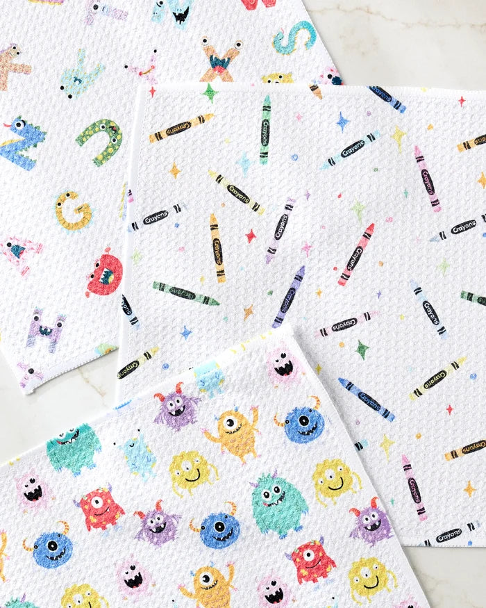 Scribble Squad by Vaishali Design Studio features three absorbent kitchen towels with playful monsters, crayons and stars, and cheerful monster faces with alphabet letters on a light marble background—adding fun to your kitchen.