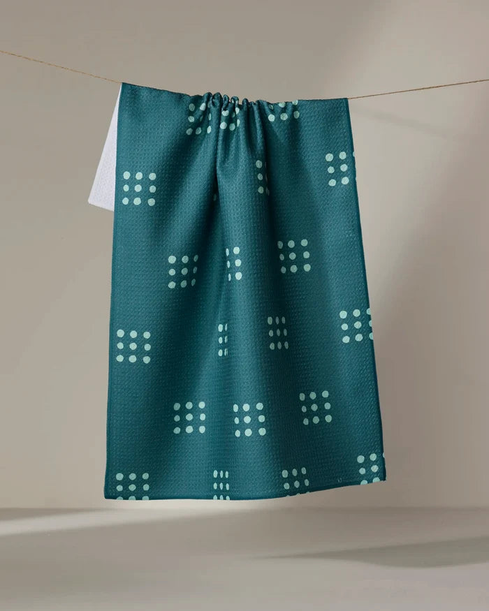 The Elizabeth Edwards Seafoam Grid tea towel, made from recycled materials, showcases a geometric light blue dot pattern and hangs on a clothesline with a neutral backdrop.