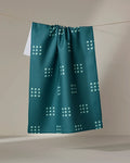 The Elizabeth Edwards Seafoam Grid tea towel, made from recycled materials, showcases a geometric light blue dot pattern and hangs on a clothesline with a neutral backdrop.