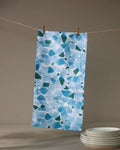 The Geometry Seaglass ultra-absorbent towel in a blue and white pattern hangs from a clothesline with wooden clothespins, above a stack of white plates on a gray surface.