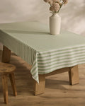 A light wooden table covered with Geometry’s Seagrass green and white stain-resistant tablecloth holds a white vase with pink flowers; a wooden stool sits nearby on the light wooden floor.