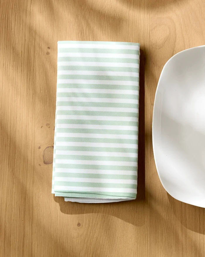 The Geometry Seagrass napkin, featuring light green and white stripes, is neatly folded on a wooden table beside a white square-edged plate, creating a warm and inviting scene.