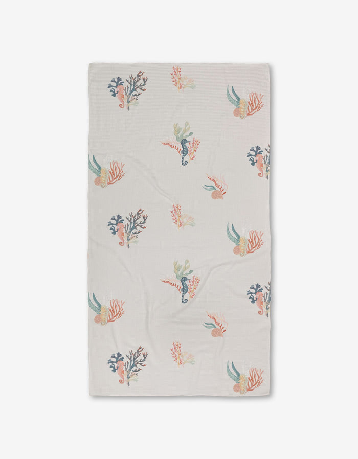 The Seahorse Serendipity Luxe Bath Towel by Janna Sue Design is a white, rectangular towel featuring colorful sea creatures, coral, and seaweed in blue, pink, orange, and green. Made with ultra-absorbent Geoweave technology. Shown flat on a white background.