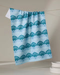 The Marta Harvey "Seashells" eco-friendly kitchen towel, featuring a blue and white scalloped fan pattern, hangs on a clothesline in a minimal room with light decor. A stack of white plates rests in the lower right corner.