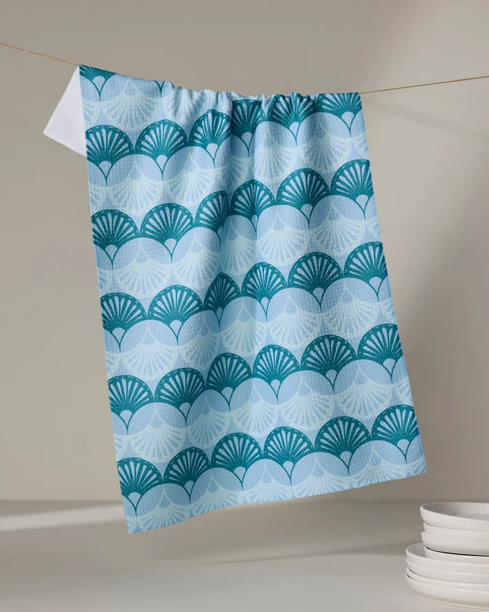 The Marta Harvey "Seashells" eco-friendly kitchen towel, featuring a blue and white scalloped fan pattern, hangs on a clothesline in a minimal room with light decor. A stack of white plates rests in the lower right corner.