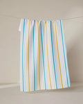 The GEOMETRY Seaside Sunshine tea towel, featuring blue and yellow vertical stripes on white, hangs on a string against a plain, light-colored background.
