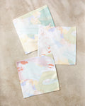The Secret Fort dishcloth set by Kristina Baker features three pastel reusable cloths made from recycled materials in soft green, yellow, peach, and blue shades with abstract patterns, arranged on a light beige surface.