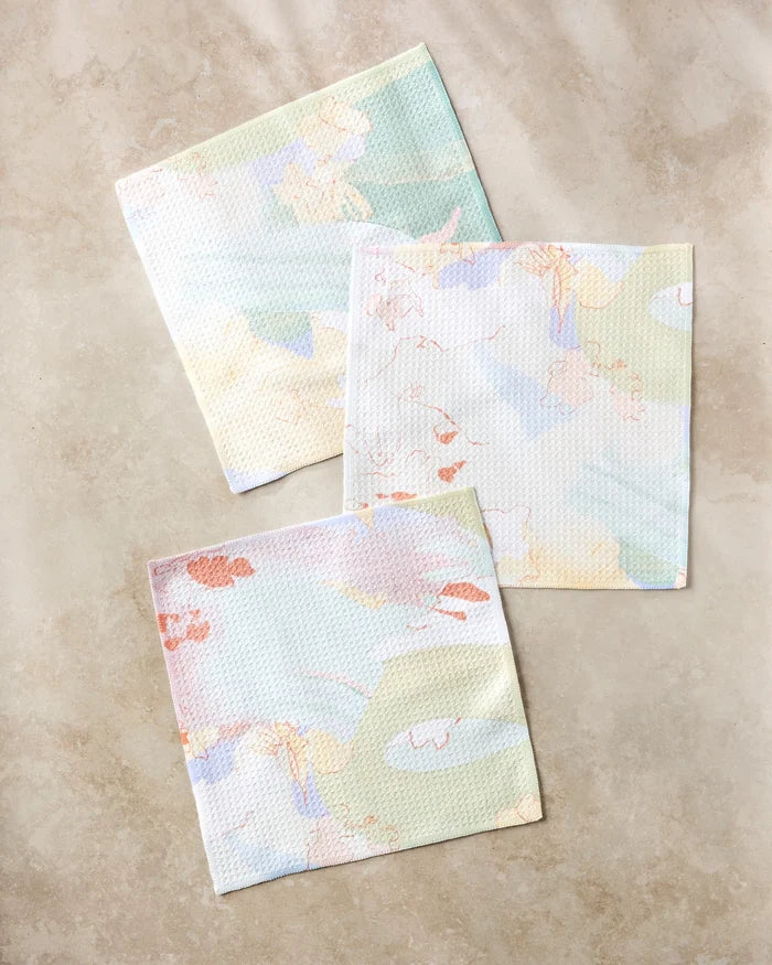 The Secret Fort dishcloth set by Kristina Baker features three pastel reusable cloths made from recycled materials in soft green, yellow, peach, and blue shades with abstract patterns, arranged on a light beige surface.