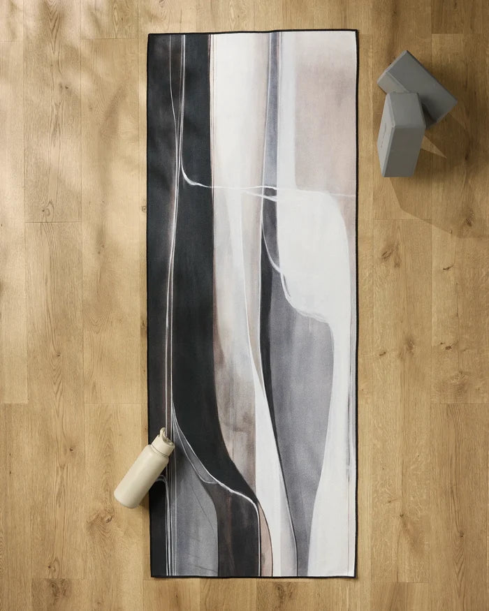 The Seduction yoga mat by Jennifer Janesko, featuring abstract black, white, and gray patterns, rests on a wooden floor alongside a beige water bottle, gray yoga blocks, and a non-slip towel for comfort and stability.