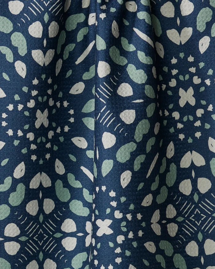 The Twig + Spark "Seeds and Nuts" kitchen towel features a dark blue eco-friendly fabric with a kaleidoscopic pattern of light green and white hearts, dots, and stars. This absorbent dish towel adds stylish charm and functionality to your kitchen.