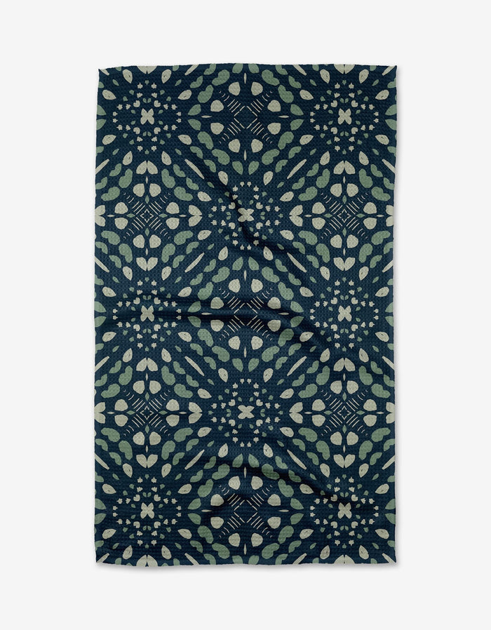 The Seeds and Nuts tea towel by Twig + Spark features a dark blue Geoweave™ fabric with light green and beige geometric leaf and petal patterns. The design is symmetrical, with the fabric showing a slightly wrinkled texture.