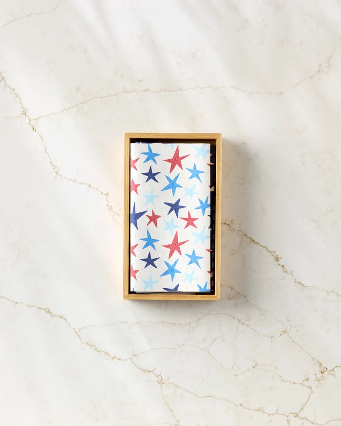 A rectangular wooden tray holds a folded white Seeing Stars kitchen towel by Mindfulnice, decorated with red and blue stars, placed on a subtly veined light marble surface.