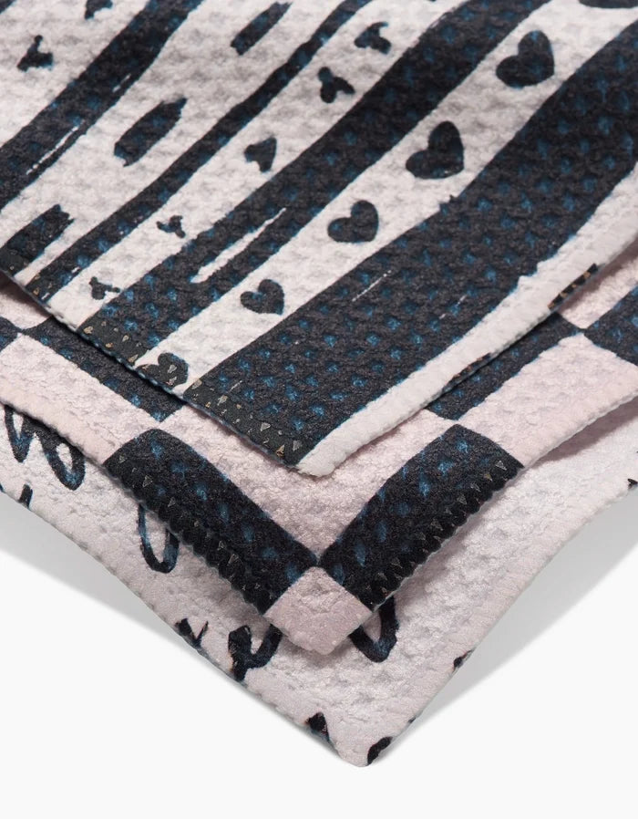 A close-up of Danelys Sidrons Self Love dishcloths shows three folded Geoweave towels with black stripes, hearts, and dots on white and pale pink, featuring visible stitching for added texture.