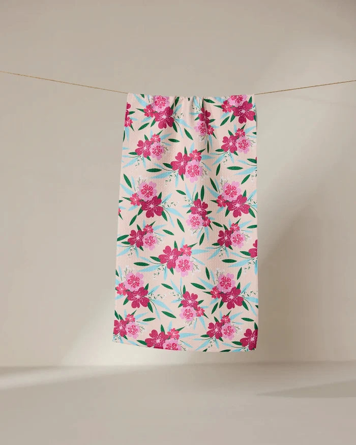 The GEOMETRY Sending Love bar towel features a white background with a bright pink floral pattern and green leaves. Made from ultra-absorbent fabric, it hangs on a clothesline against a neutral beige backdrop. Perfect for daily use.