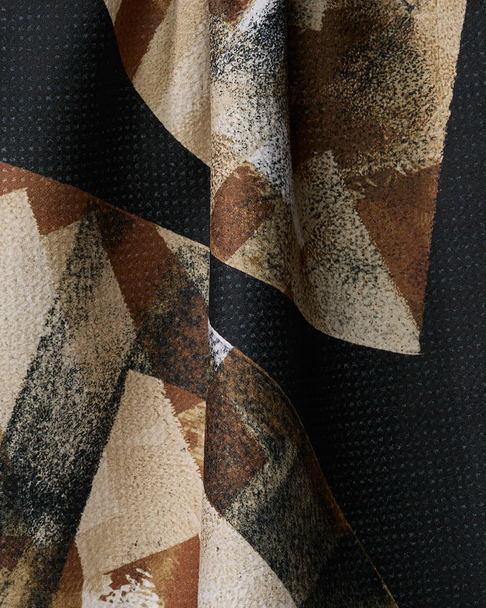 A close-up of Sepia Shift fabric by Skye Walker, featuring recycled material with abstract geometric patterns in black, beige, brown, and cream. Overlapping shapes and brush-like textures make it perfect for a stylish tea towel; the fabric appears draped.