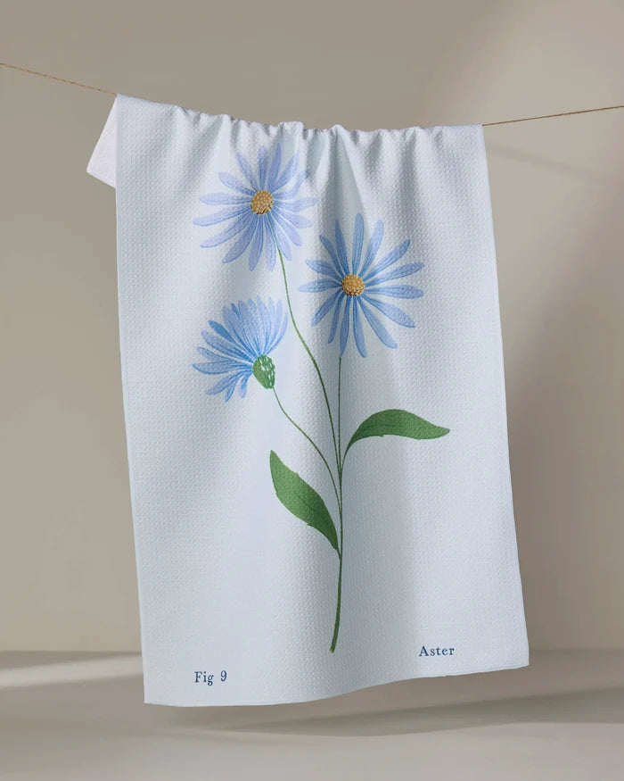 A light-colored kitchen tea towel by Linda Fox, named "September Aster," hangs on a line. It features three blue asters with green leaves, is made from recycled materials, and displays "Fig 9" and "Aster" at the bottom.