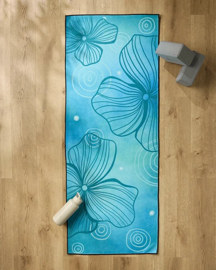 The Blissful Flamingo Serenity yoga mat, featuring abstract flower and swirl patterns, rests on a wooden floor with a white water bottle, two gray yoga blocks, and a non-slip towel for comfort and stability during practice.