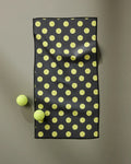 The Geometry Serve mat, a black rectangular mat made from recycled materials and patterned with yellow dots, rests on a gray surface with two yellow tennis balls placed beside it.