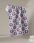 The Seventies Retro Flowers recycled kitchen towel by Claire Iglesias, featuring a blue, red, and white geometric floral pattern, hangs above a stack of white plates against a neutral background.