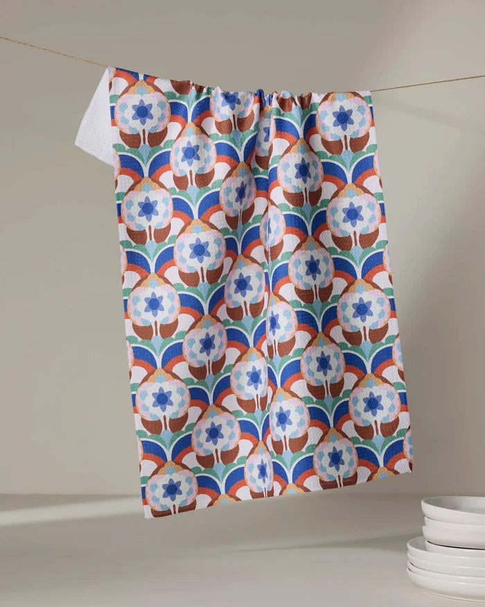 The Seventies Retro Flowers recycled kitchen towel by Claire Iglesias, featuring a blue, red, and white geometric floral pattern, hangs above a stack of white plates against a neutral background.