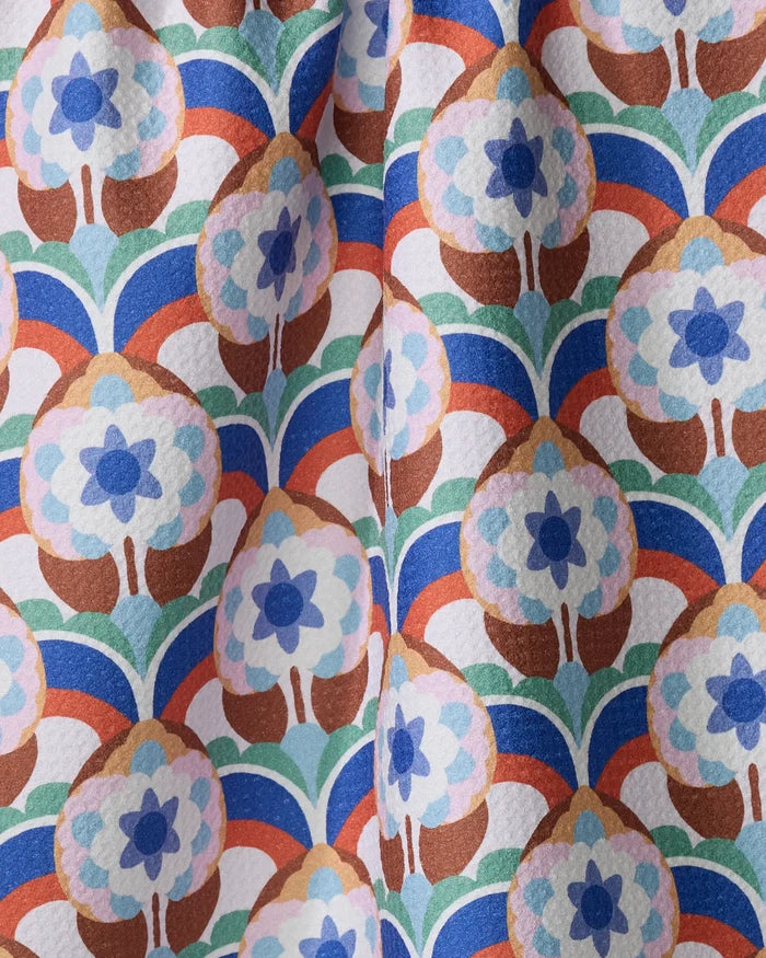 The Seventies Retro Flowers tea towel by Claire Iglesias showcases rows of stylized florals in blue, brown, green, and orange with scalloped accents on white. Quick-drying fabric features slight folds for a vibrant retro look.