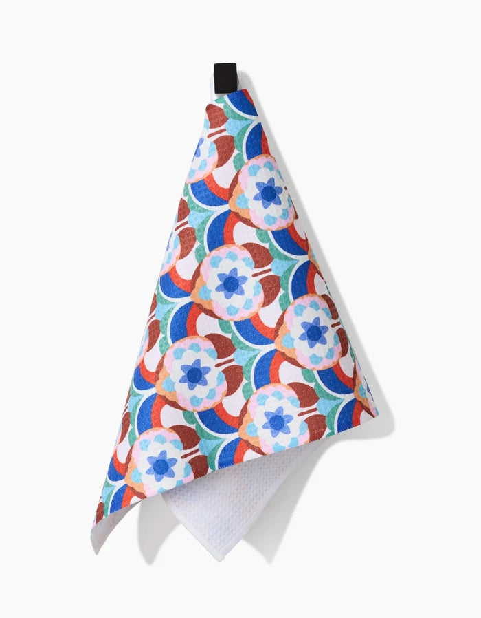 Claire Iglesias’ Seventies Retro Flowers bar towel, made from recycled materials, displays a vibrant blue, orange, red, and green geometric floral pattern and hangs from a black loop against a white background.
