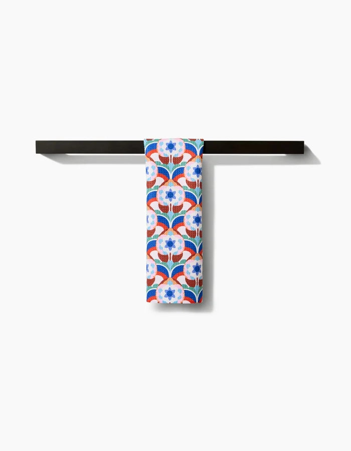 The Seventies Retro Flowers bar towel by Claire Iglesias, featuring a blue, red, and white floral design on soft Geoweave fabric, is neatly folded over a simple black bar against a plain white background.