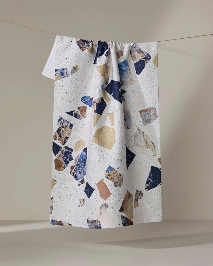 The Geometry Shale tea towel, made from recycled materials, features an abstract blue, beige, and brown terrazzo pattern. It hangs on a string against a plain beige background.