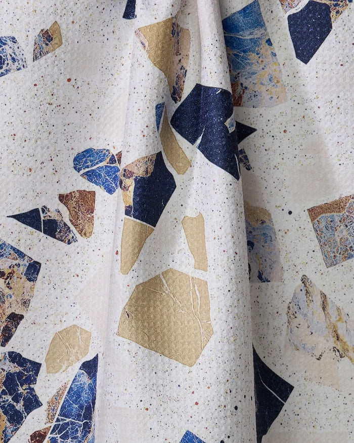 A close-up of Geometry’s Shale tea towel shows a terrazzo-inspired pattern with irregular blue, gold, and beige shapes on an off-white background with colorful speckles. The ultra-absorbent towel is slightly gathered in the center.