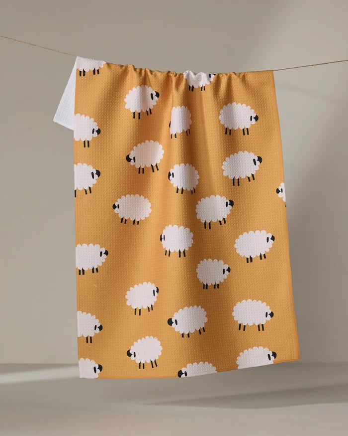 A yellow "Sheep" kitchen tea towel by Jennifer Bouron, featuring a pattern of white cartoon sheep, hangs on a clothesline against a neutral background.