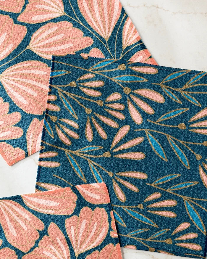 Three "Sheila Flower" fabric swatches by Amy MacCready, with pink flowers and green leaves on blue backgrounds and gold accents, are arranged on marble—ideal for eco-friendly kitchen towels.