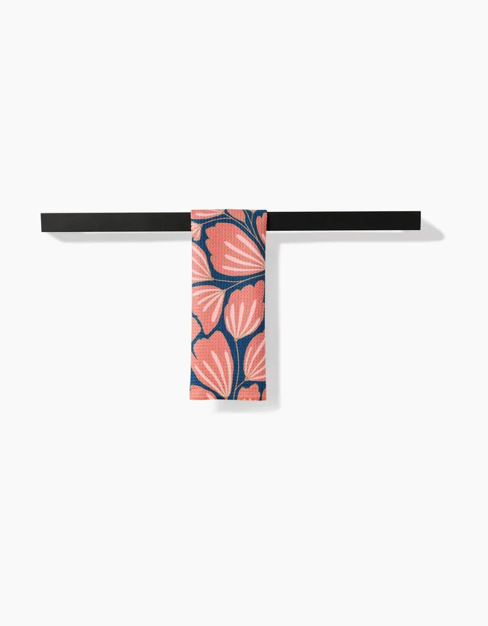 A black horizontal towel rack holds the Sheila bar towel by Amy MacCready, featuring a vibrant pink and coral floral pattern on dark blue Geoweave fabric, set against a plain white wall.