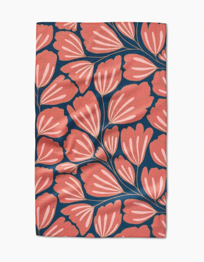The Sheila tea towel by Amy MacCready showcases large stylized coral-pink leaves with white accents on dark blue, made from recycled materials and shown laid flat with slight wrinkles.