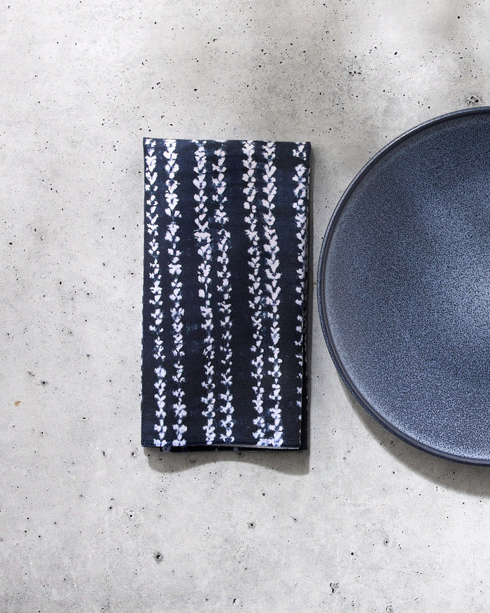 The GEOMETRY Shibo reusable table napkin, featuring a white floral pattern, is neatly folded beside a dark gray ceramic plate on a light gray textured surface.