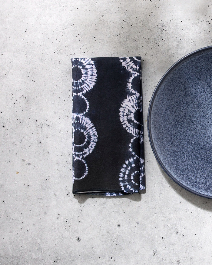 The Shibo napkin by GEOMETRY, featuring a black reusable fabric with a white circular tie-dye pattern, is displayed next to a dark blue plate on a light gray concrete surface.