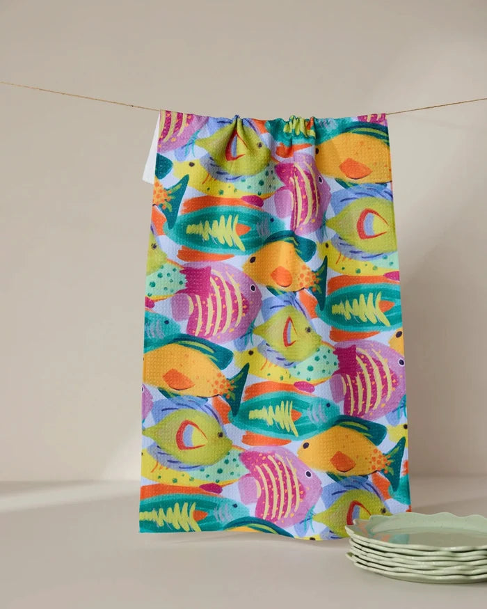 The Geometry Shoaling eco-friendly kitchen towel, featuring a vibrant fish pattern, hangs from a string against a neutral backdrop with green plates stacked nearby.