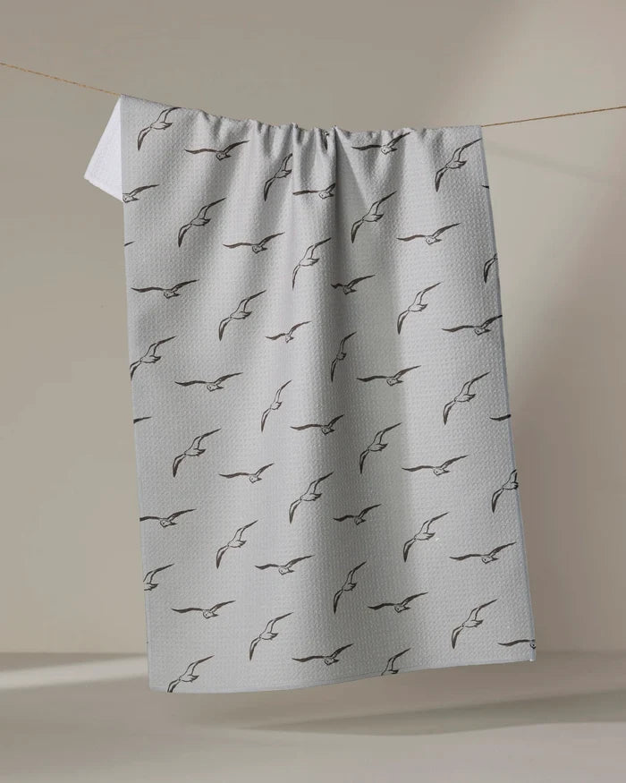 The Jenny Keller Shoreline Scout is a light gray absorbent dish towel featuring a pattern of simple black outlined birds in flight, shown hanging neatly from a clothesline against a neutral background.